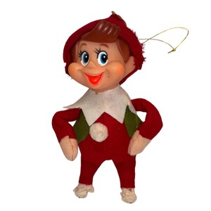 Puede incluir: Un adorno de elfo vintage con cara de plástico moldeado, ojos azules pintados y boca roja. El cuerpo es de fieltro rojo con detalles en verde, un cuello de fieltro blanco y un pompón blanco en el pecho. Incluye un cordón dorado para colgar.
