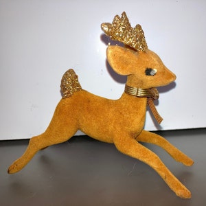 Pode incluir: Uma figura de rena de veludo marrom com chifres de glitter dourado e uma fita dourada em volta do pesco&ccedil;o.