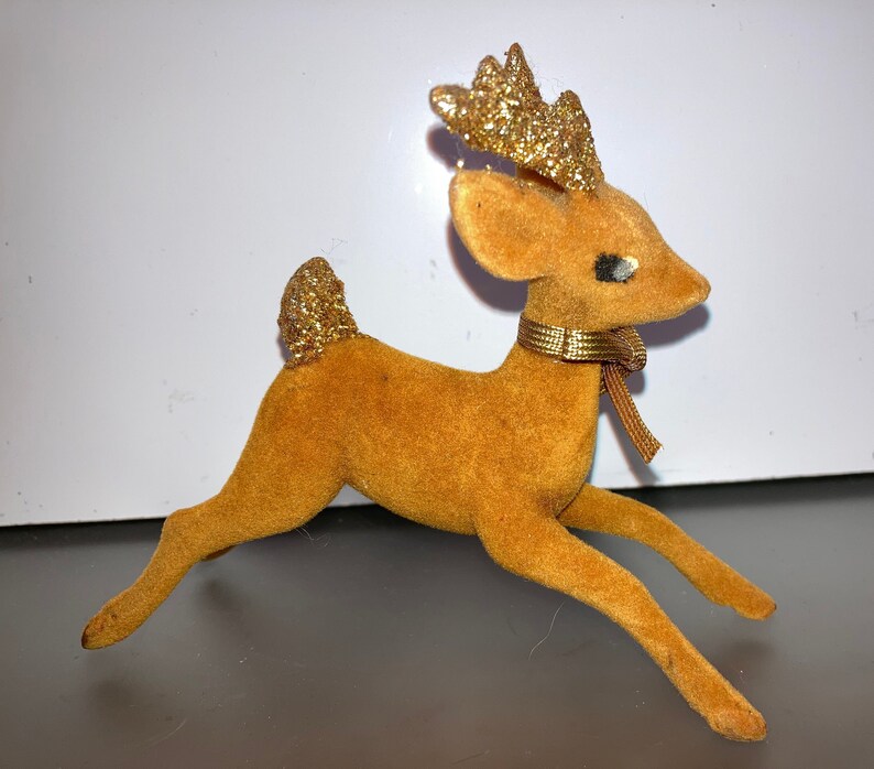Pode incluir: Uma figura de rena de veludo marrom com chifres de glitter dourado e uma fita dourada em volta do pesco&ccedil;o.