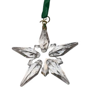 May include: A clear, faceted crystal star ornament with five points. It hangs from a gold-colored metal loop and a green ribbon. The star is designed to catch and reflect light, creating a sparkling effect.