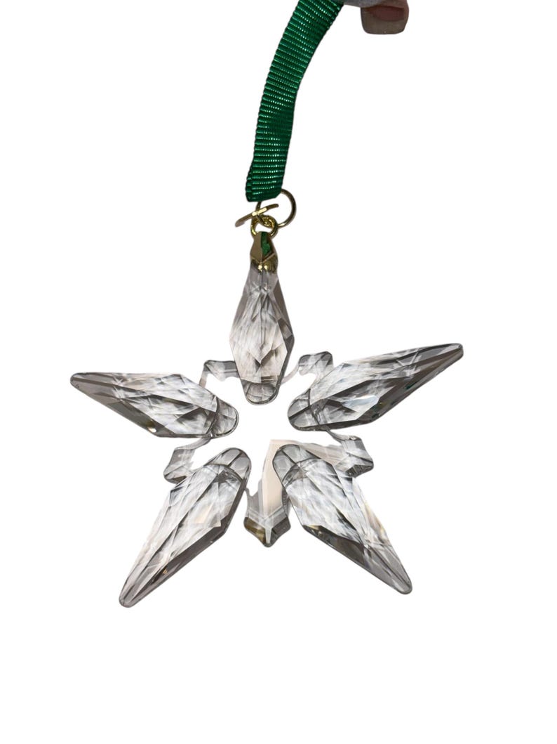 May include: A clear, faceted crystal star ornament with five points. It hangs from a gold-colored metal loop and a green ribbon. The star is designed to catch and reflect light, creating a sparkling effect.