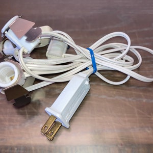 May include: White electrical cord with three white light bulb sockets and a white plug. The cord is coiled and has a blue rubber band holding it together.