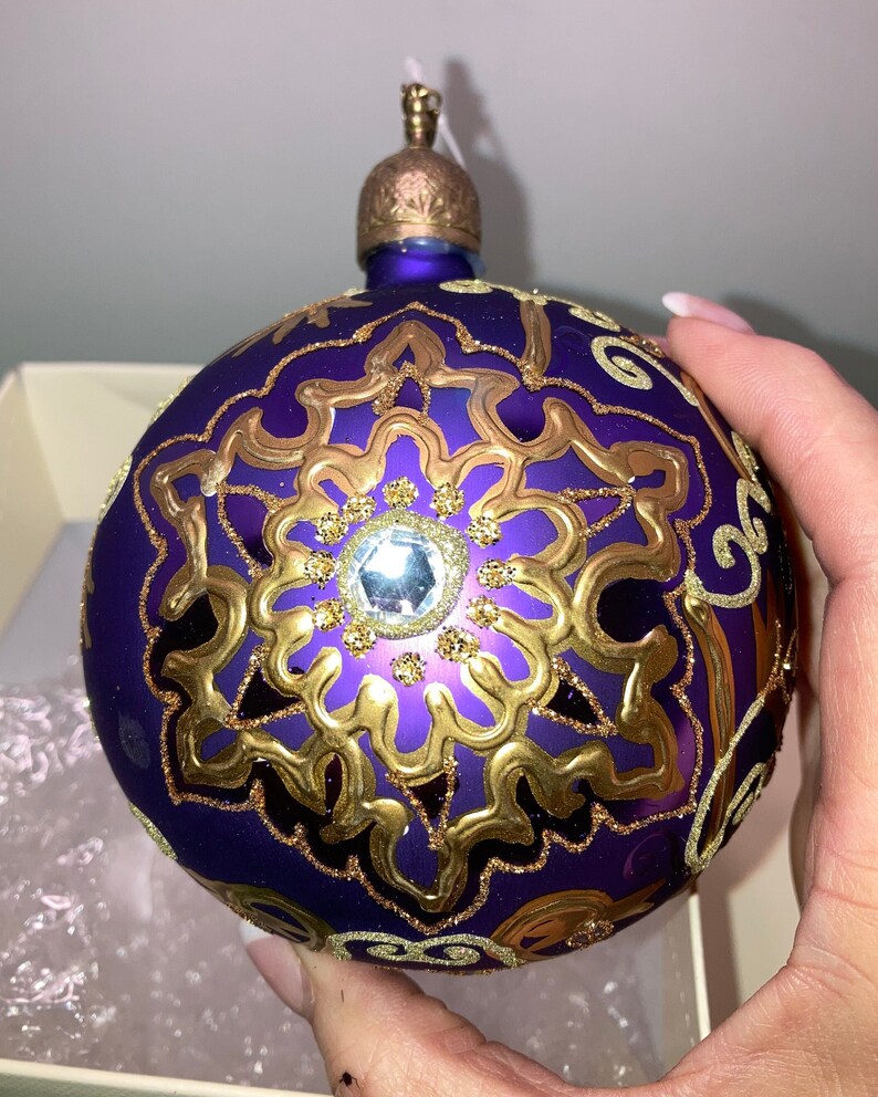 May include: Purple glass Christmas ornament with gold glitter and a gold snowflake design. The ornament has a clear crystal in the center of the snowflake.