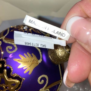 May include: Purple glass Christmas ornament with gold detailing and a gold hanging loop. The ornament has a sticker that says "SKU 8015 9468" and another sticker that says "MADE IN POLAND".