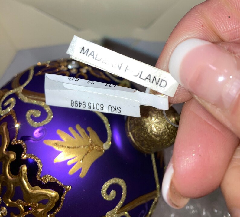 May include: Purple glass Christmas ornament with gold detailing and a gold hanging loop. The ornament has a sticker that says "SKU 8015 9468" and another sticker that says "MADE IN POLAND".