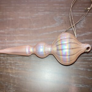 May include: A pink iridescent glass Christmas tree topper with a bulbous top and a long, pointed bottom. The topper has a string loop for hanging.