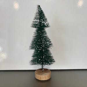 May include: A miniature artificial Christmas tree with dark green needles and a dusting of white snow. It stands on a round, natural wood base. This decorative tree is perfect for the festive season.