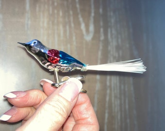 Può includere: Ornamento vintage a forma di uccello in vetro con corpo argentato, dettagli blu e rossi e una lunga coda bianca. L'uccello è attaccato a una clip argentata, probabilmente per fissarlo a un ramo. Una classica decorazione natalizia.