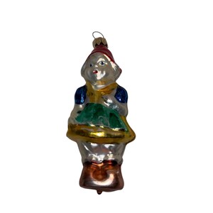 May include: Vintage Christmas ornament in the shape of a figure holding a basket. The ornament features a red hat, blue sleeves, and a gold-colored collar and basket. The figure is holding a green object.