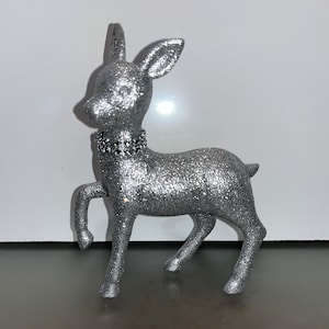 Puede incluir: Una figurita de ciervo de purpurina plateada con un collar de pedrería negra. El ciervo está de pie sobre una superficie blanca.