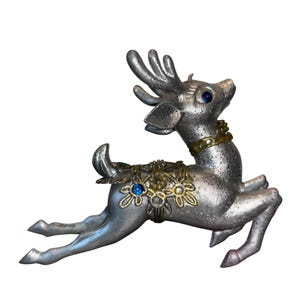Op de afbeelding: Zilverkleurig rendierbeeldje met blauwe edelsteenogen en accenten. Het rendier heeft een goudkleurige kraag en bloemversieringen op de rug. Het beeldje staat in een sprong, met uitgestrekte poten.