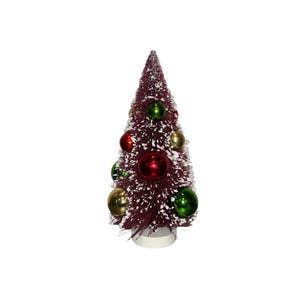 Op de afbeelding: Een kleine, bordeauxrode kerstboom van flessenborstel met witte flocking. De boom is versierd met rode, groene en gouden ballen. De boom staat op een witte basis.