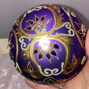 May include: Purple glass Christmas ornament with gold and glitter accents. The ornament features a floral design with intricate details.