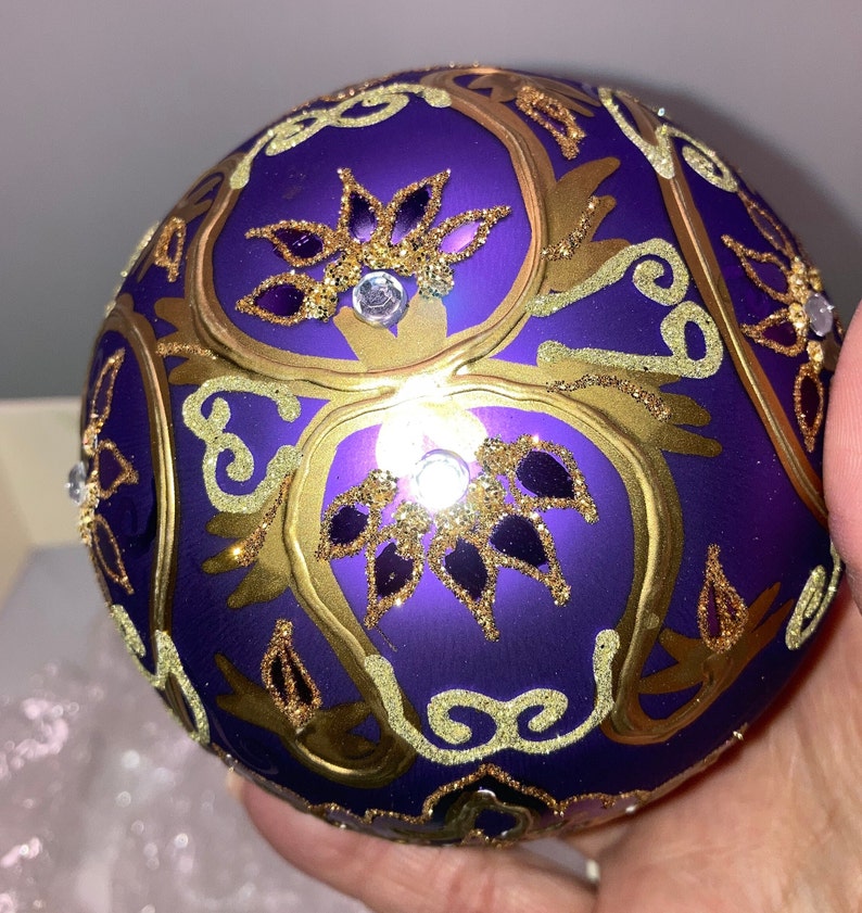 May include: Purple glass Christmas ornament with gold and glitter accents. The ornament features a floral design with intricate details.