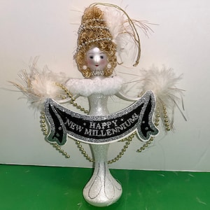 Könnte beinhalten: Ein weißer Glas-Anhänger in Form einer Frau, die einen weißen Pelzkragen und eine goldene Krone trägt. Sie hält ein schwarzes Banner mit der Aufschrift "Happy New Millenium".
