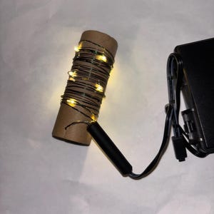 Pode incluir: Um conjunto de luzes de fadas brancas quentes enroladas em torno de um tubo de papelão, conectadas a uma bateria preta com um fio.