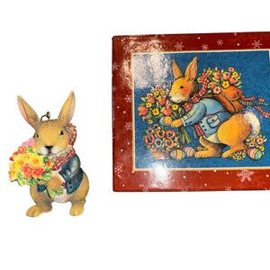 May include: A decorative Easter bunny figurine holding a bouquet of yellow and pink flowers, wearing a blue jacket and a red scarf. Beside it is a red box with a painting of a rabbit with flowers and Easter eggs.