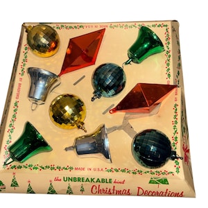 Op de afbeelding: Een vintage doos met kerstversiering. De doos bevat verschillende vormen, waaronder facetgeslepen bollen, bellen en ruitvormen, in goud, groen, zilver en rood. De doos is bruin met groene en rode accenten en de tekst "the UNBREAKABLE kind Christmas Decorations."