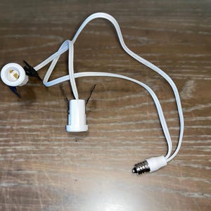 May include: Two white electrical cord ends with a white wire connecting them. The cord ends have a standard light bulb socket.