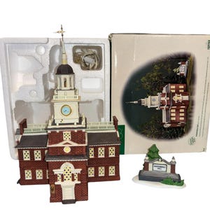 May include: A miniature replica of Independence Hall, featuring a brick facade, white trim, and a clock tower with a gold spire. The model is displayed in its original packaging, alongside a small Liberty Bell sign.