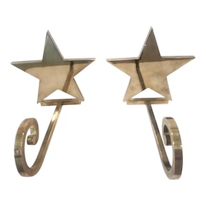 May include: A pair of polished brass wall hooks, each featuring a five-pointed star design at the top and a curved, scroll-shaped hook extending downward. The metallic surface has a reflective, smooth finish with clean, sharp edges throughout.