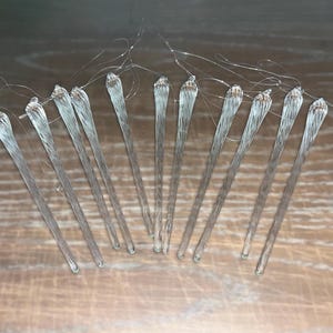 Puede incluir: Un conjunto de 12 adornos de cristal transparente en forma de carámbano con un diseño retorcido. Los adornos miden aproximadamente 15 cm de largo y tienen un lazo de alambre plateado en la parte superior para colgar.
