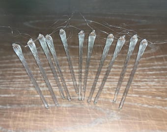 May include: A set of 12 clear glass icicle ornaments with a twisted design. The ornaments are approximately 6 inches long and have a silver wire loop at the top for hanging.