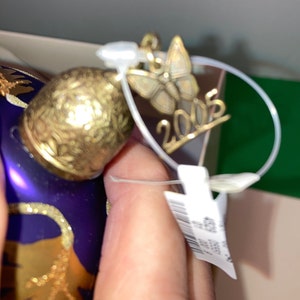 May include: A purple glass Christmas ornament with gold accents and a butterfly charm. The ornament has the year 2005 written on it.
