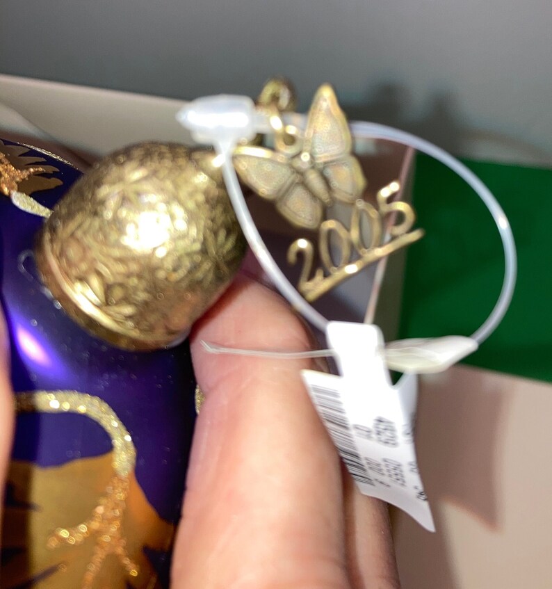 May include: A purple glass Christmas ornament with gold accents and a butterfly charm. The ornament has the year 2005 written on it.