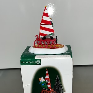 Puede incluir: Un velero rojo y blanco con un trineo rojo y una figura de una persona con un atuendo verde y blanco. La figura sostiene una cuerda roja atada a la parte superior del velero. La figura está de pie en el trineo. El trineo está sobre una base blanca. La base tiene las palabras "SAVA" impresas en ella. La base está sobre una caja verde con la palabra "DEPARTMENT" impresa en ella.