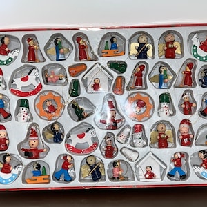 May include: A set of 36 wooden Christmas ornaments in a clear plastic box. The ornaments are painted in various colors, including red, green, blue, yellow, and white. The ornaments feature a variety of designs, including angels, stars, snowmen, and rocking horses.