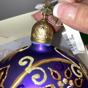 May include: Purple glass Christmas ornament with gold accents and a butterfly topper. The ornament has a gold and purple design with glitter accents.