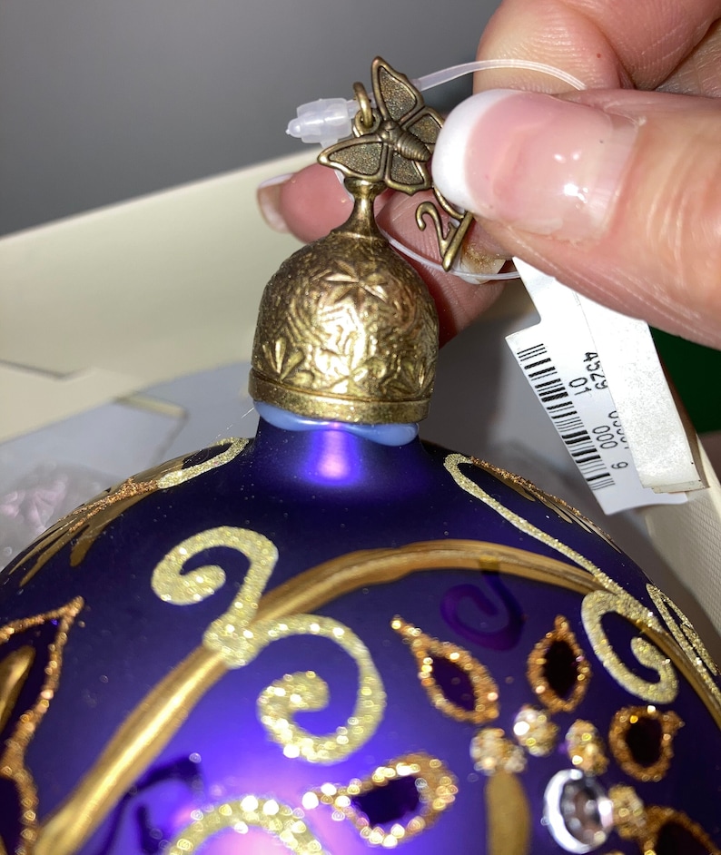 May include: Purple glass Christmas ornament with gold accents and a butterfly topper. The ornament has a gold and purple design with glitter accents.