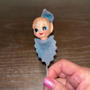 Puede incluir: Un adorno de cabeza de elfo de fieltro azul y blanco con ojos azules y nariz roja. El elfo está unido a un tallo de alambre blanco.