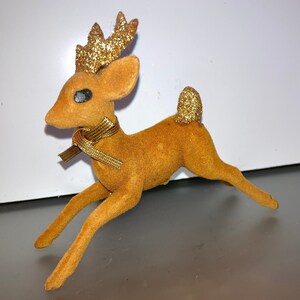 Pode incluir: Uma figura de rena de veludo marrom com chifres de glitter dourado e uma fita dourada em volta do pesco&ccedil;o. A rena est&aacute; em uma pose de corrida com a cabe&ccedil;a virada para o lado.