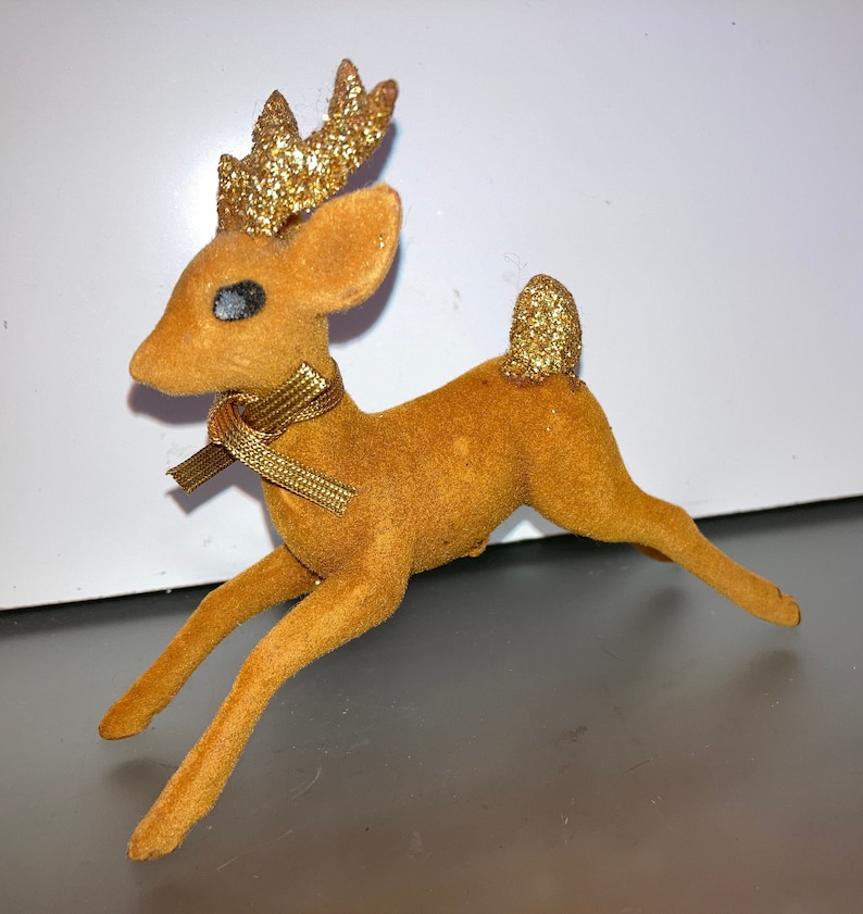 Pode incluir: Uma figura de rena de veludo marrom com chifres de glitter dourado e uma fita dourada em volta do pesco&ccedil;o. A rena est&aacute; em uma pose de corrida com a cabe&ccedil;a virada para o lado.
