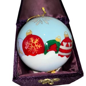 May include: A Christmas bauble with a light blue backdrop, decorated with red, green, and white festive designs. The bauble is presented in a purple velvet box, with a gold hanging string.