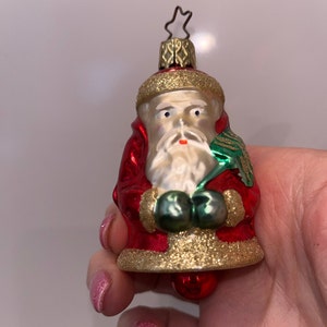 May include: A vintage Christmas ornament shaped like Santa Claus. The ornament features a red coat with gold glitter trim, a white beard, and green mittens. A small red ball hangs at the bottom, and a gold star tops the ornament.