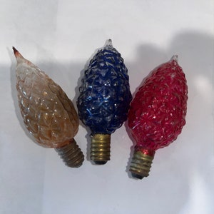 May include: Three vintage Christmas light bulbs shaped like pine cones. The bulbs are clear glass with a textured surface. One bulb is clear, one is blue, and one is red.