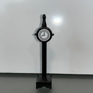 May include: A miniature black clock tower with a round clock face. The clock face is white with black Roman numerals and hands. The tower has a rectangular base and a decorative top.