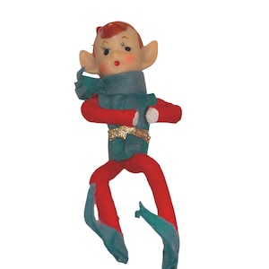 Peut inclure: Une figurine de lutin articulée avec un visage pâle, des traits peints, des cheveux roux et des oreilles pointues. Le lutin porte un gilet et une écharpe verts, une ceinture dorée à paillettes, des manches rouges à revers blancs et des collants rouges avec des chaussures pointues vertes.