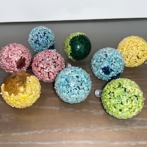 May include: Assortment of eight small, round, glass baubles covered in sparkly glitter. The baubles are in various colours, including blue, green, yellow, pink, and light blue.