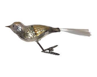 May include: A vintage-style glass bird ornament with a reflective silver and gold finish. The bird features a pointed beak, a textured body, and a long, thin white fibre tail. It is attached to a metal clip base for secure placement on a branch.