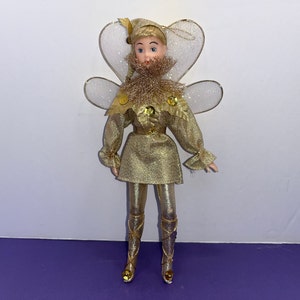 May include: A gold fairy doll with a white glitter wings, a gold glitter dress, and a gold glitter crown. The doll has a white face with painted blue eyes and a pink mouth.