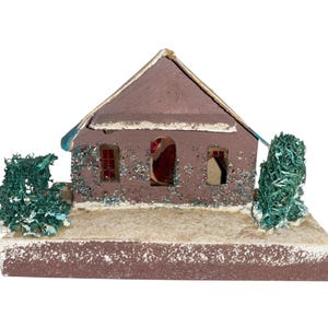 Puede incluir: Una casa marrón en miniatura con tejado a dos aguas, dos ventanas y una puerta arqueada. La casa está decorada con purpurina y tiene dos arbustos verdes texturizados a cada lado. La base es de color marrón claro.