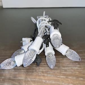 Puede incluir: Una cadena de luces navideñas blancas con bombillas transparentes. Las luces están enchufadas y listas para usar.