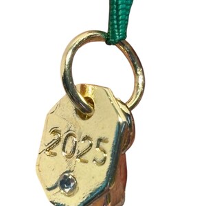 May include: A gold-colored charm with the year "2025" engraved on it, along with a small, clear gem. The charm is attached to a gold ring and a green ribbon. The charm has a geometric shape.