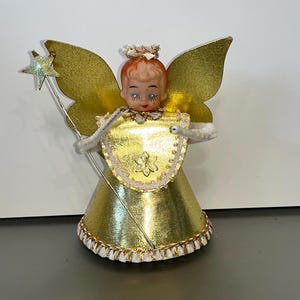 May include: A gold angel tree topper with a star wand, white wings, and a white and gold dress. The angel has a plastic face with blue eyes and a red and white crown.