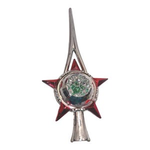 May include: A silver-toned tree topper featuring a central star design with red points and a circular centre filled with small, reflective green and silver mosaic-like pieces. The ornament has a tall, pointed silver spire and a conical base.