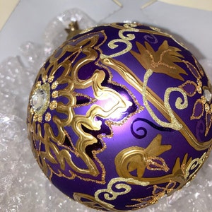 May include: Purple Christmas ornament with gold and glitter accents. The ornament features a floral design.